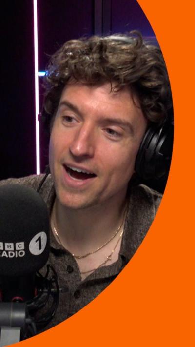 A man with short, curly brown hair speaks into a microphone