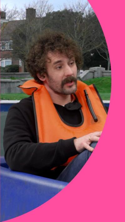 Josh Pugh has dark curly hair and a moustache and is wearing an orange life vest whilst sitting in a pedalo.