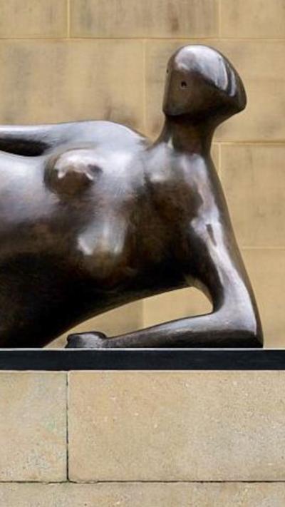 A bronze statue on a plinth. The figure is reclining in a relaxed manner. The wall behind is light stone.