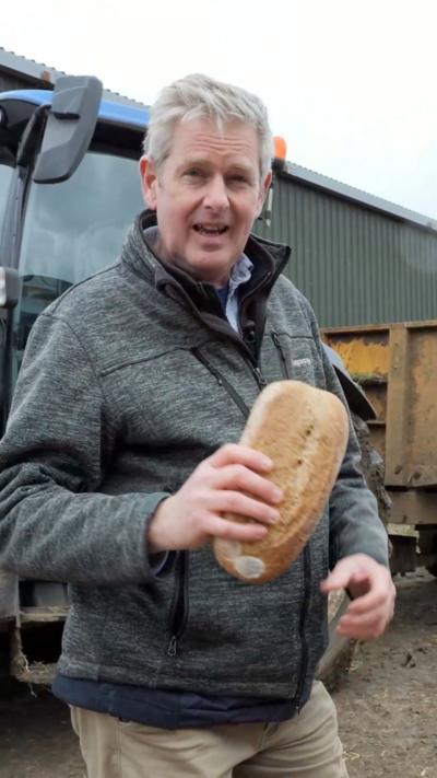 A man holding a baguette whilst standing in front of a tractor on a farm.