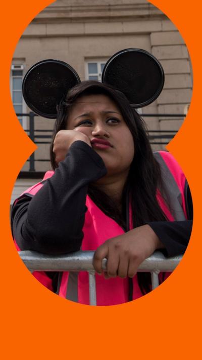 A woman with Mickey Mouse ears and a high vis jacket on leans against a metal railing looking fed up