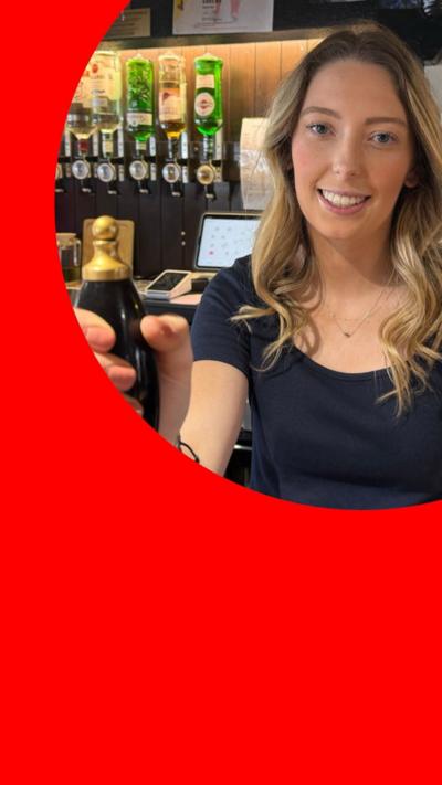 A woman, Neve, stands behind the bar in a pub. There are bottles of spirits and good luck cards behind her