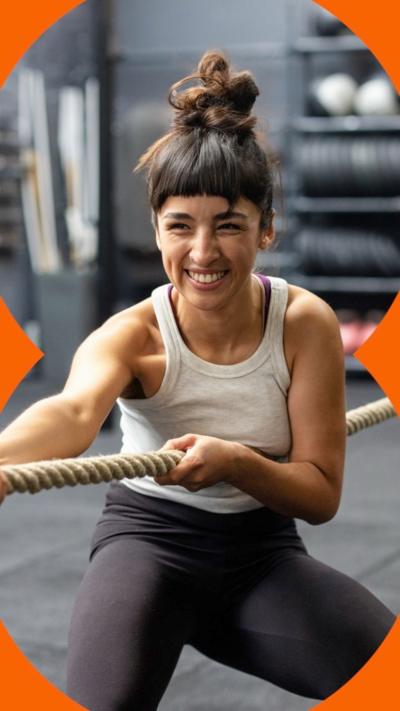 A woman smiles, with her hair in messy top knot, playing tug of war at a training gym.