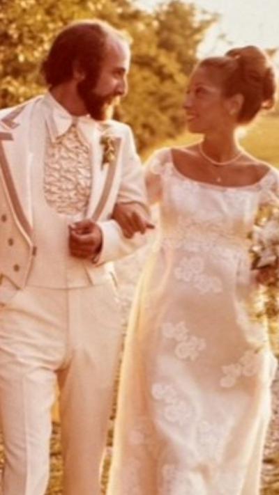 A man and a woman are dressed in all white 1970s era wedding clothes walking arm in arm next to each other. 