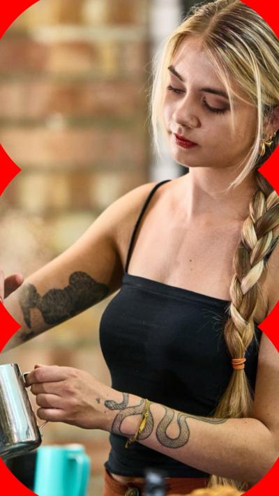 A young barista with blonde hair and tattoos serves coffee in a cafe.