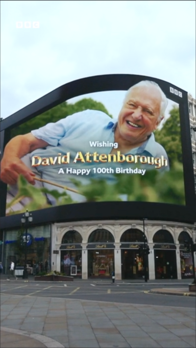 An image of David Attenborough on the Piccadilly Circus screen in central London with text saying 'wishing David Attenborough a happy 100th birthday' as people watch from the street