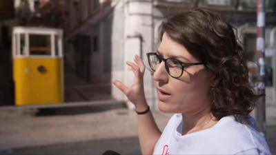 Rasha Abdo wearing a white top and black rimmed glasses gestures towards the undamaged yellow and white funicular carriage.