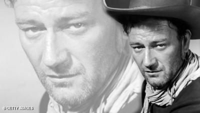 Black and white image of John Wayne is pictured in costume from the 1948 film '3 Godfathers', directed by John Ford. He is on the right hand side of the picture, head and shoulders, showing his scraf and hat. In the background on the left is an enlarged image of his face from the same image, slightly faded into the background.