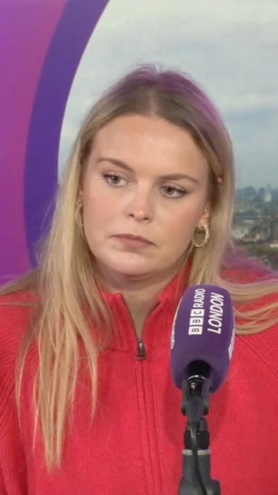 A woman in a red sweater stands in front of a cityscape backdrop behind a microphone with "BBC Radio London" branding.
