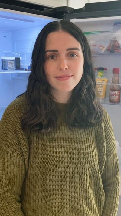 A lady with long brown, wavy hair smiles at the camera as she stands in front of an open fridge wearing a green jumper.
