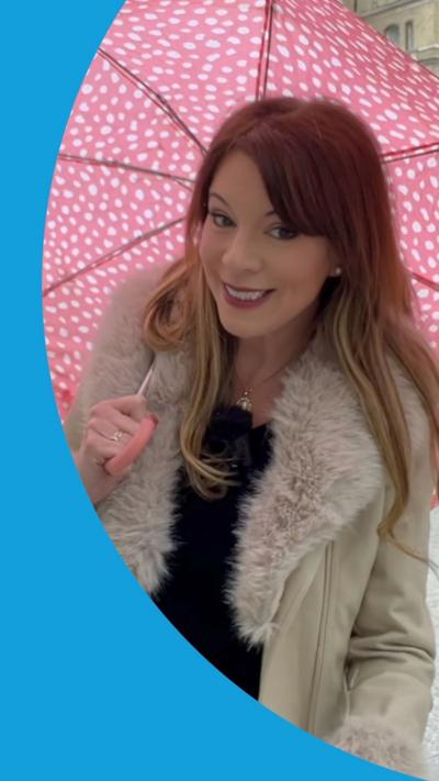 Woman with red hair, wearing a cream coat and holding a red umbrella with white spots.