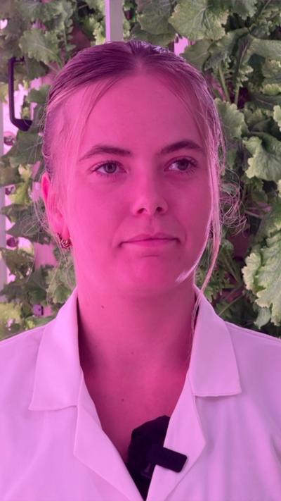 Laura Nelson from Innovation Agritech Group wearing a white lab coat, while inside a vertical farm with plants behind her.
