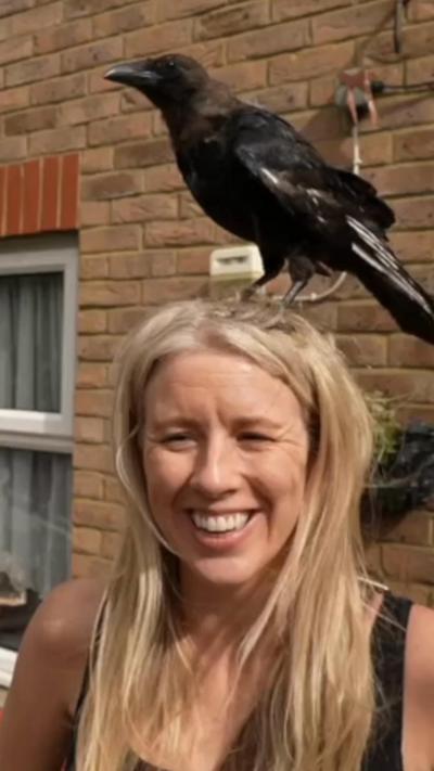 A woman smiles as a crow sits on her head. She is wearing a black vest top and is standing in front outside a brick house. 