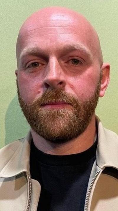 A bald man with beard stood against a light green studio wall. He is wearing a camel coloured jacket and black top