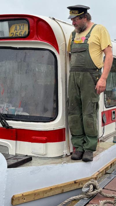 Man wearing dungarees is standing on a boat made from an old bus 