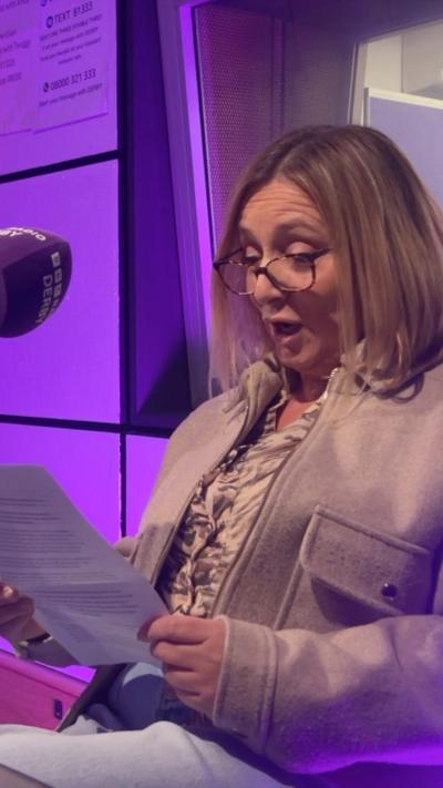 A person seated in what appears to be a radio or recording studio. She's holding an A4 sheet of paper and wearing a light-coloured jacket over a patterned top. She appears to be reading what's on the piece of paper. A large, purple studio microphone with visible branding is positioned to the left. 