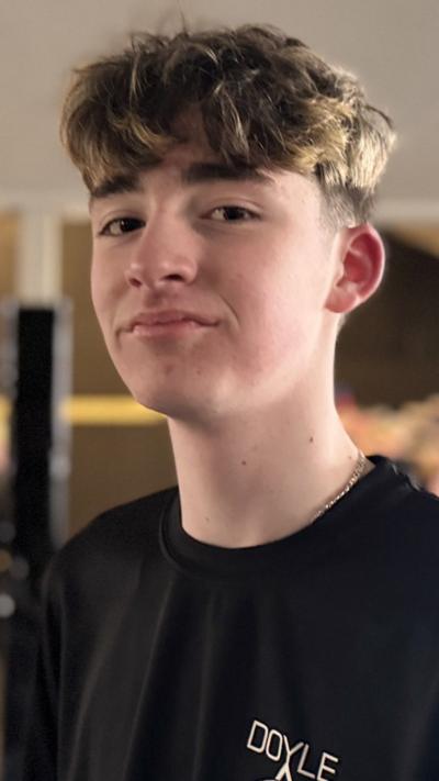 A boy with brown hair is turned sideways smiling into the camera, he is dressed in a black t-shirt. Behind him is an out-of-focus martial arts studio.