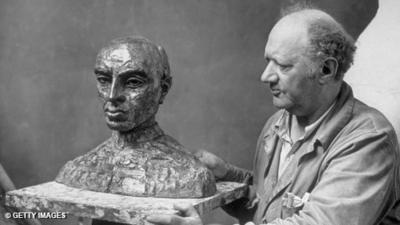 Black and white. Jacob Epstein on right of frame, in work overalls, holding onto and looking at a sculpture of a bust of a bald man.