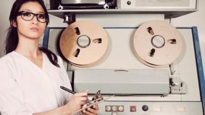 Image of a lady wearing white with a clipboard, stood in front of a reel to reel machine