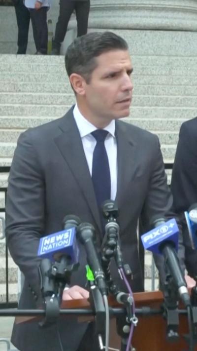 A man wearing a grey suit talks behind microphones.