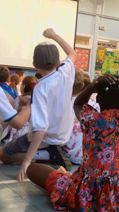 Crowds of children, some with white and red England shirts, have their backs turned to the camera, watching the England game on a big screen