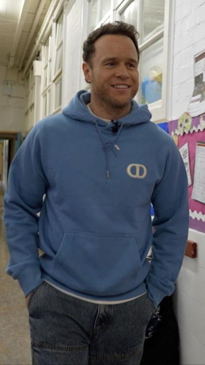 Olly Murs in a blue hoodie walks down a school corridor, beside colourful bulletin boards displaying educational materials.