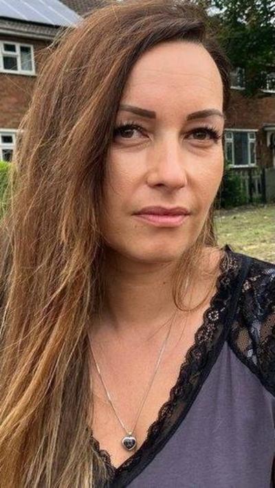 A woman with long brown hair, swept to one side, sat on a bench. She is wearing a short sleeved top with black lace shoulders and purple front