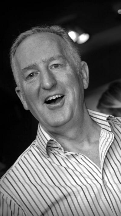 A black and white image of a man with short grey hair smiling. He's wearing an open necked striped shirt