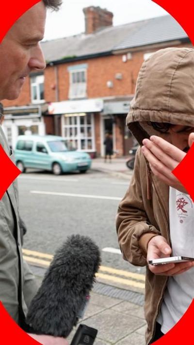 A man in a hooded jacket covers his face while being asked questions by journalist Ed Thomas in the street