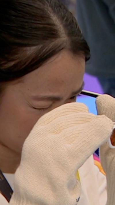 A woman with dark hair and white top with blue, purple and orange stripes on the sleeves tries out gloves and glasses intended to show what life is like for a dementia patient