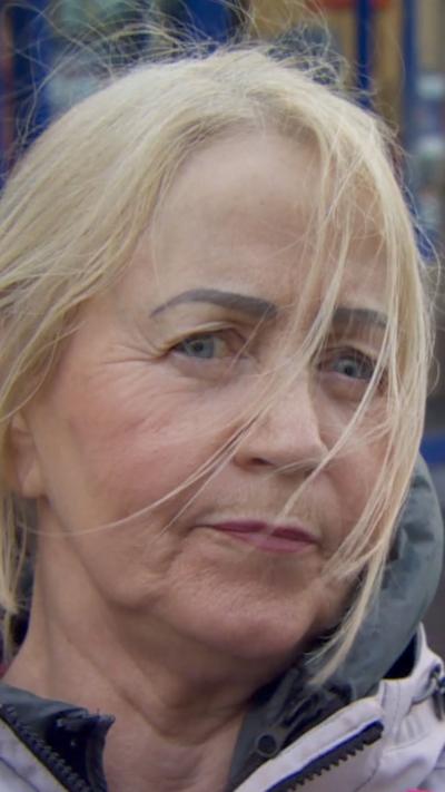 A woman with blonde hair looks at the camera. She has pink lipstick and a pink coat. Her hair is blown slightly across her face.