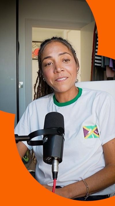 A woman, Miquita Oliver, wears a white T-shirt with the Jamaica flag and has long brown hair. She sits behind a microphone