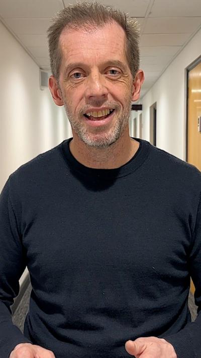 A white man with brown hair in a black shirt looking at the audience.