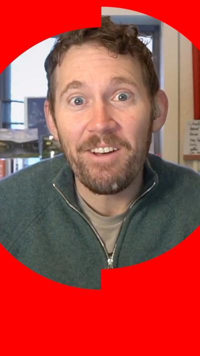 A man with short brown hair, a moustache and beard talks to the camera. He is wearing a green jumper with an unzipped collar.