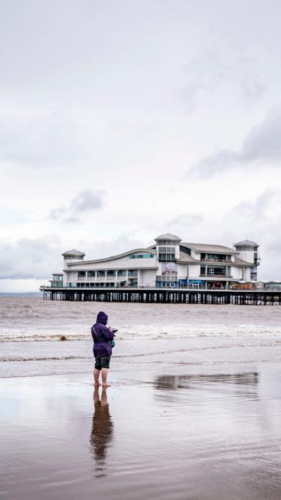 A woman stood on the shore, bare foot, at Weston beach. She has on a dark purple hoodie and rolled up jeans. The pier is stretching along the shoreline in the distance. 