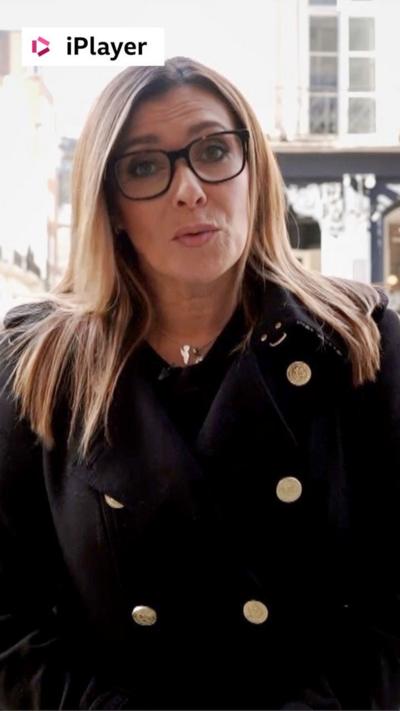 A woman, Kym Marsh stands on a street. She has long hair and wears a black jacket and glasses.