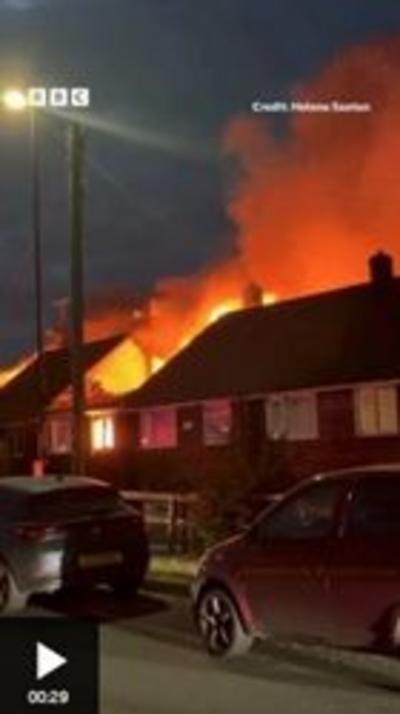 Picture of a street where several houses are on fire. Flames and smoke can be seen behind the houses, and there are parked cars in the foreground