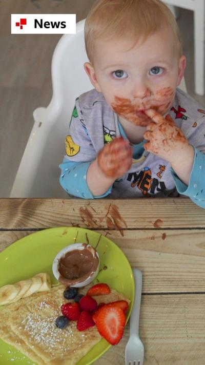 A toddler with his face covered in chocolate sits in a highchair and looks up at the camera. A plate with pancakes, fruit and chocolate sits in front of him.