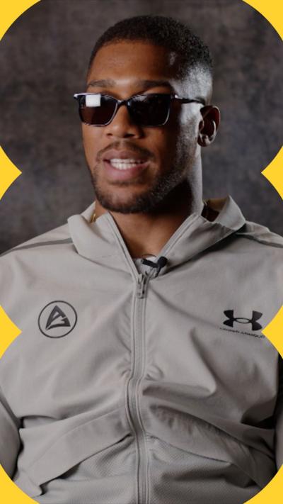 Anthony Joshua is wearing a zipped up, stone-coloured top. He has cropped black hair and is wearing shades.