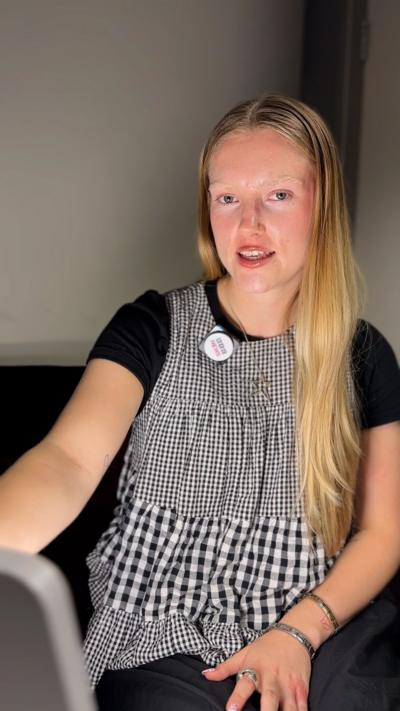 A young woman with long blond hair wearing a black T-shirt, and a black-and-white checkered dress. She is sitting and next to her is a bright, rectangular light therapy lamp. The light is illuminating her face.