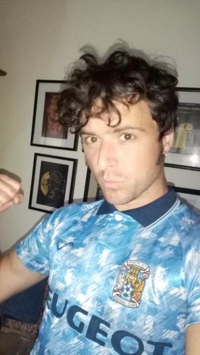 A man with brown curly hair wearing a 1990s Coventry City sky blue football shirt