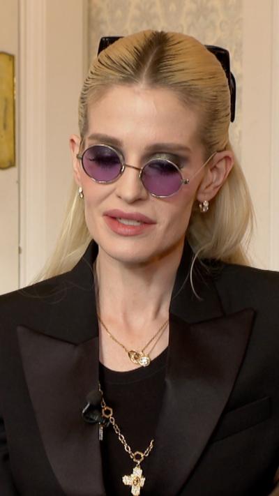 Woman with swept back blond hair, tinted glasses and black suit