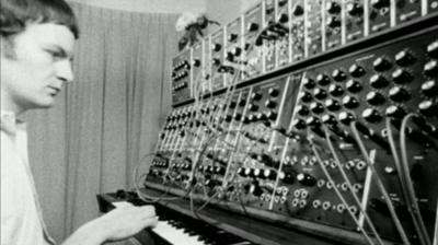 A man, sitting down, plays a Moog Synthesizer