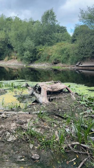 A rusted, rotten car overgrown with weeds and surrounded by algae sits in the water of the River Lagan. The wheels are submerged beneath the water but the body of the car is entirely visible. The windows are down. It's a dusty orange-pink colour.