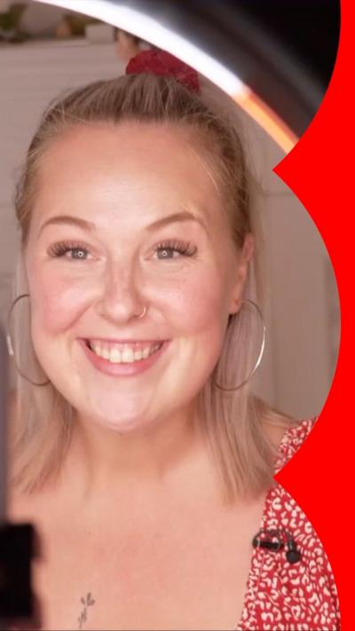 A woman smiles while looking through a ring light. She has long blonde hair and wears a red patterned dress with hoop earrings 