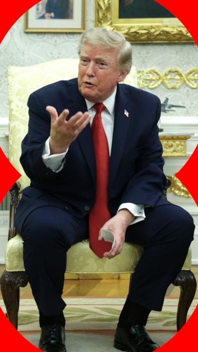 A blonde man gesticulates while sat in a cream chair. He is wearing a blue suit and red tie.