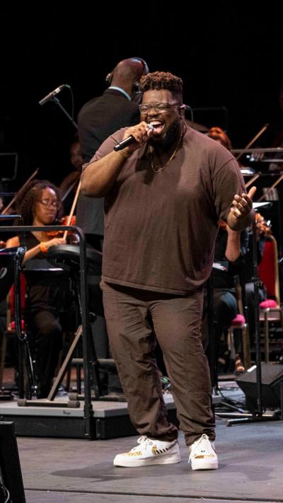 A smiling male vocalist in a brown t-shirt and glasses sings into a handheld mic on stage, with orchestra musicians seated behind him