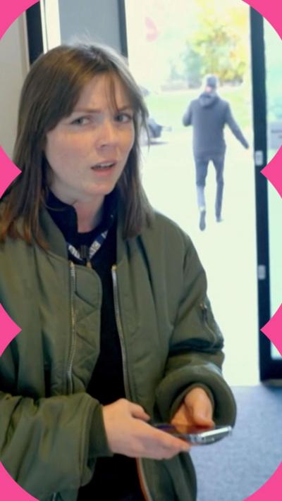 A woman stands and looks confused. She is wearing a green jacket and holding a phone.