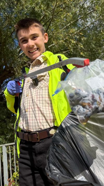 Young boy with brown hair, wearing a smart plaid shirt and yellow hi-vis smiles to camera holding a bag and litter picker.