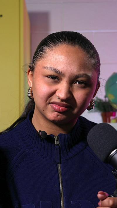 A young woman wearing a dark zip‑up sweater speaks into a studio microphone, in a softly lit background.